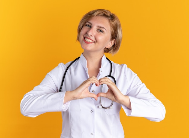 Foto grátis jovem médica sorridente, vestindo bata médica com estetoscópio, mostrando um gesto de coração isolado em um fundo laranja