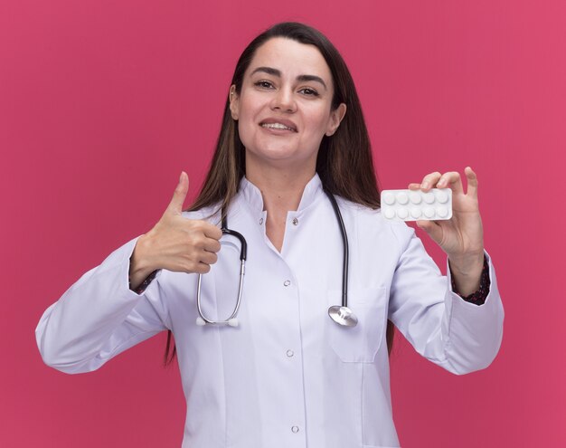 Jovem médica sorridente, vestindo túnica médica com estetoscópio, segurando o pacote de remédios e os polegares para cima, isolados na parede rosa com espaço de cópia