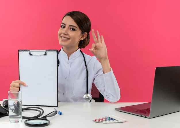 Jovem médica sorridente, vestindo túnica médica com estetoscópio, sentada na mesa de trabalho no computador com ferramentas médicas, segurando a prancheta e mostrando o gesto certo na parede rosa