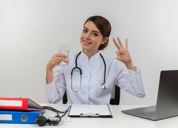 Jovem médica sorridente, vestindo túnica médica com estetoscópio sentada na mesa de trabalho no computador com ferramentas médicas, segurando um copo de água e mostrando o gesto certo na parede branca