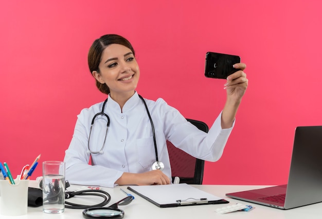 Jovem médica sorridente vestindo túnica médica com estetoscópio sentada na mesa de trabalho no computador com ferramentas médicas, tire uma selfie com espaço de cópia