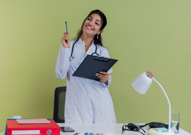 Jovem médica sorridente vestindo túnica médica e estetoscópio atrás da mesa com ferramentas médicas, segurando a prancheta e a caneta isoladas