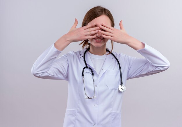 Jovem médica sorridente, vestindo túnica médica e estetoscópio, fechando os olhos com as mãos na parede branca isolada