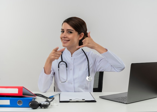 Jovem médica sorridente, vestindo túnica médica e estetoscópio, sentada à mesa com ferramentas médicas e laptop, fazendo um gesto de chamada, apontando isolado na parede branca