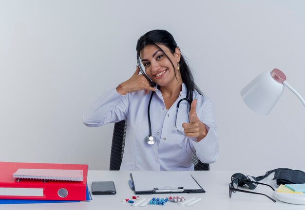 Jovem médica sorridente, vestindo túnica médica e estetoscópio, sentada à mesa com ferramentas médicas, olhando para fazer o gesto de chamada e o gesto de pistola isolado