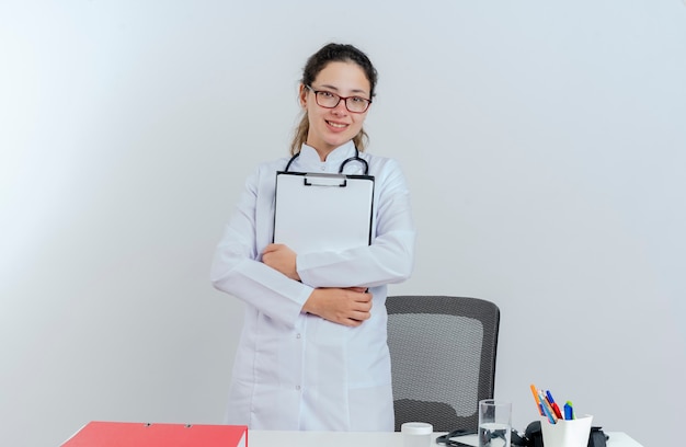 Jovem médica sorridente, vestindo túnica médica, estetoscópio e óculos, em pé atrás da mesa com ferramentas médicas, segurando a prancheta isolada