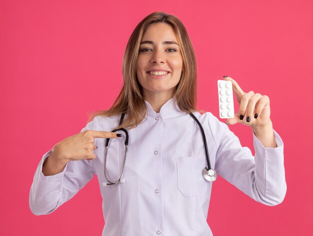 Jovem médica sorridente, vestindo túnica médica, segurando um estetoscópio e apontando para pílulas isoladas na parede rosa