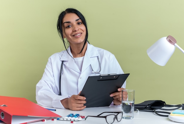 Jovem médica sorridente, vestindo um manto médico com estetoscópio, sentada na mesa com ferramentas médicas, segurando uma prancheta isolada sobre fundo verde oliva