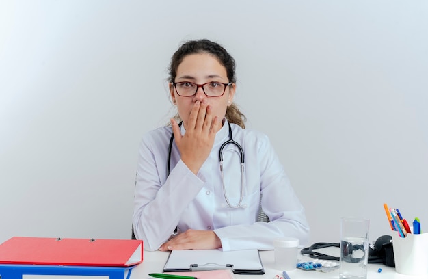 Jovem médica surpreendida usando túnica médica, estetoscópio e óculos, sentada na mesa com ferramentas médicas, olhando colocando a mão na boca isolada