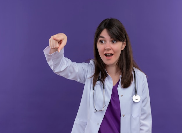 Jovem médica surpresa com roupão médico e estetoscópio apontando para o lado em fundo violeta isolado com espaço de cópia