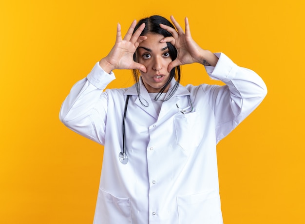 Jovem médica surpresa, vestindo bata médica com estetoscópio, abrindo os olhos com as mãos isoladas em um fundo amarelo