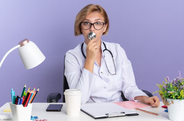 Jovem médica surpresa, vestindo túnica médica com estetoscópio e óculos, sentada à mesa com ferramentas médicas isoladas sobre fundo azul