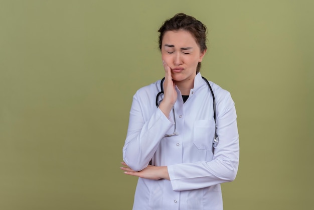 Jovem médica triste, usando bata médica e estetoscópio, colocando a mão no dente dolorido na parede verde