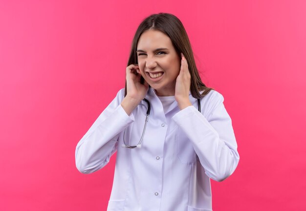 jovem médica usando estetoscópio bata médica com as orelhas fechadas na parede rosa isolada