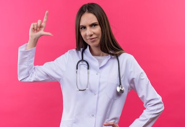 Foto grátis jovem médica usando estetoscópio bata médica mostrando o tamanho na parede rosa isolada