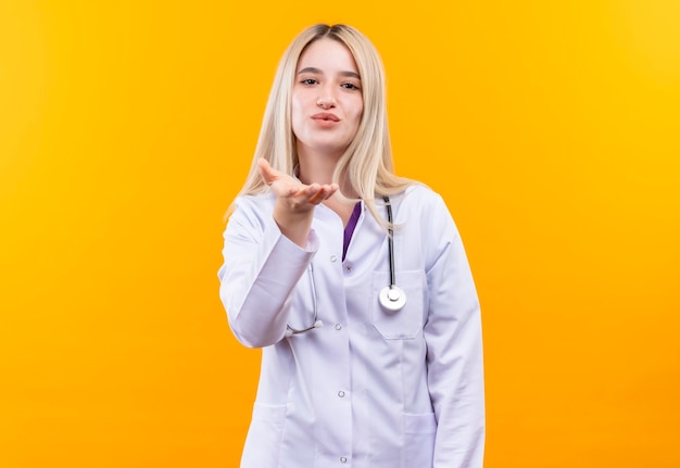 Jovem médica usando estetoscópio e vestido de médico, estendendo a mão para a câmera na parede amarela isolada