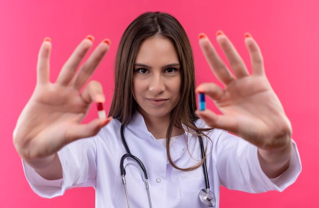 jovem médica usando estetoscópio jaleco segurando comprimidos para a câmera na parede rosa isolada