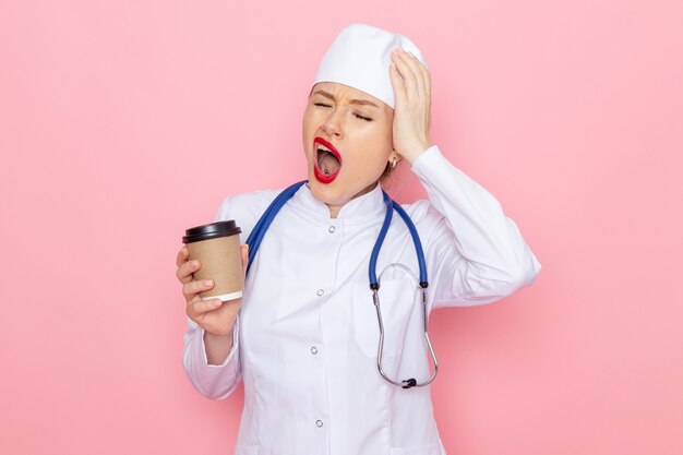 Jovem médica vestida de terno branco com estetoscópio azul segurando uma xícara de café de plástico no hospital médico de medicina espacial rosa