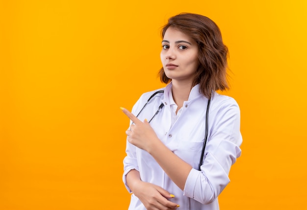 Jovem médica vestindo jaleco branco com estetoscópio apontando com o dedo indicador para o lado e rosto sério em pé sobre uma parede laranja