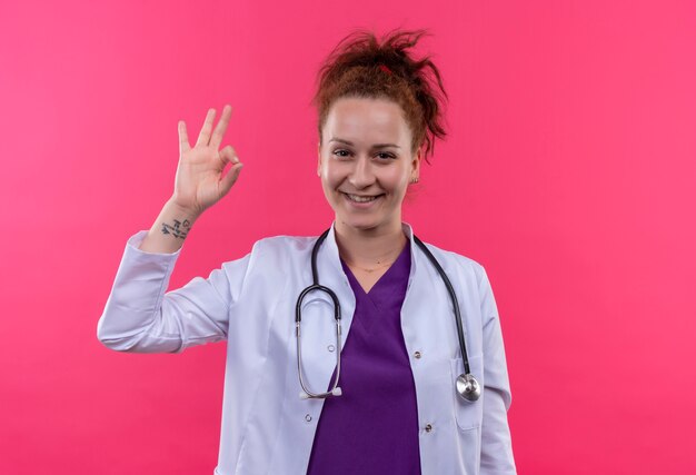 Jovem médica vestindo jaleco branco com estetoscópio e sorrindo com uma cara feliz fazendo sinal de ok em pé sobre a parede rosa