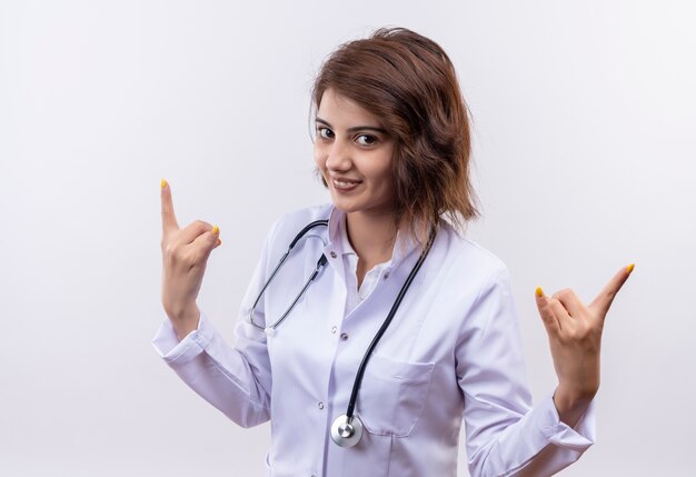 Jovem médica vestindo jaleco branco com estetoscópio fazendo símbolos de rock com dedos sorrindo alegremente em pé sobre uma parede branca