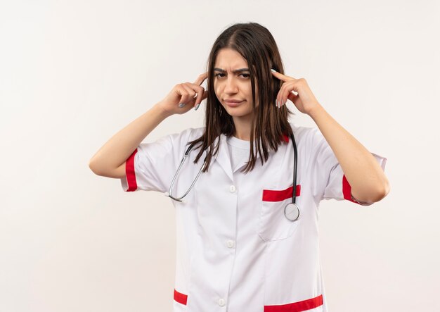 Jovem médica vestindo jaleco branco com estetoscópio no pescoço apontando para as têmporas, parecendo cansada e entediada em pé sobre uma parede branca