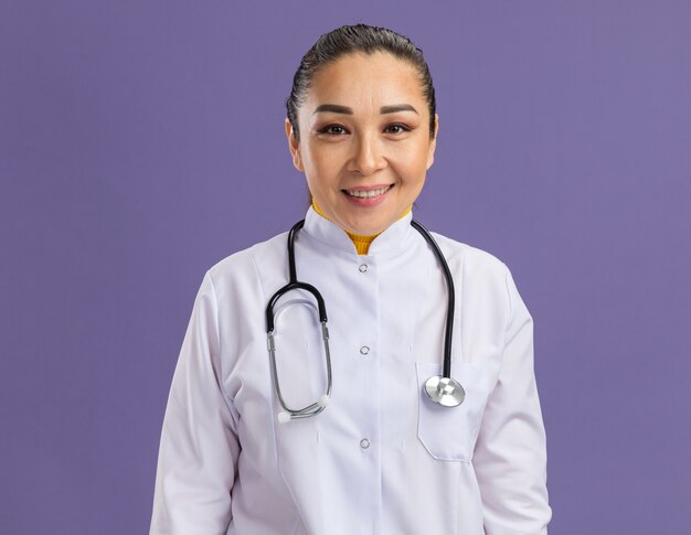 Jovem médica vestindo jaleco branco com estetoscópio no pescoço e sorrindo confiante em pé sobre a parede roxa