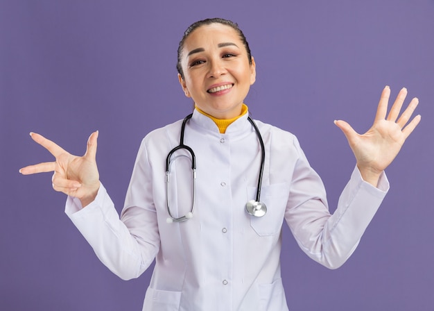 Jovem médica vestindo jaleco branco com estetoscópio no pescoço e sorrindo mostrando o número oito com os dedos em pé sobre a parede roxa