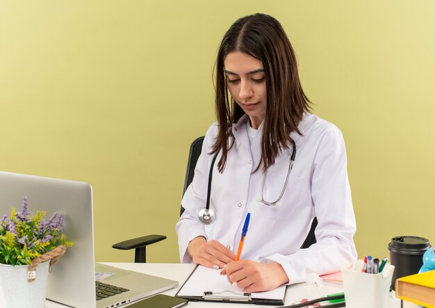 Jovem médica vestindo jaleco branco com estetoscópio no pescoço escrevendo algo com cara séria, sentada à mesa com laptop sobre parede de luz