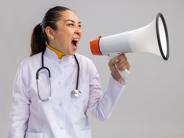 Jovem médica vestindo jaleco branco com estetoscópio no pescoço gritando para o megafone com expressão agressiva