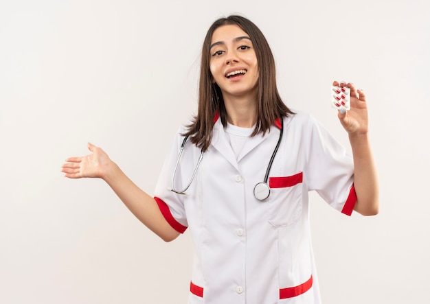 Jovem médica vestindo jaleco branco com estetoscópio no pescoço mostrando bolha com comprimidos apontando com o braço da mão para o lado feliz e surpresa em pé sobre uma parede branca