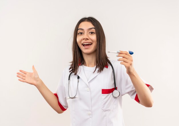 Jovem médica vestindo jaleco branco com estetoscópio no pescoço segurando um termômetro digital, feliz e animada, apontando com o braço da mão para o lado, parada sobre uma parede branca