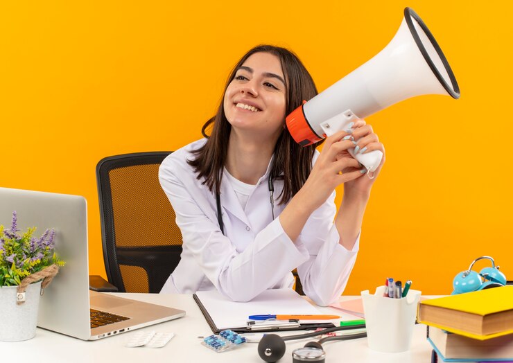 Foto grátis jovem médica vestindo jaleco branco com estetoscópio segurando um megafone e sorrindo com uma carinha feliz sentada à mesa com o laptop e documentos na parede laranja