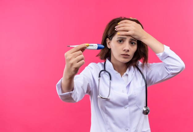 Jovem médica vestindo jaleco branco com estetoscópio segurando um termômetro digital em choque tocando a cabeça