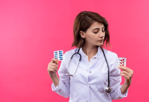 Foto grátis jovem médica vestindo jaleco branco com estetoscópio segurando uma bolha com comprimidos olhando para ela com cara séria