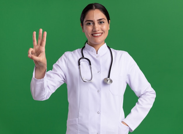 Jovem médica vestindo jaleco com estetoscópio olhando para frente, mostrando e apontando para cima com os dedos número três em pé sobre a parede verde