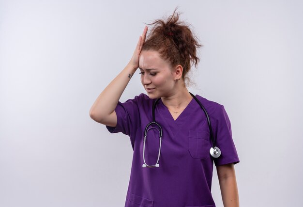 Jovem médica vestindo uniforme médico com estetoscópio parecendo confusa tocando sua cabeça por engano em pé sobre uma parede branca