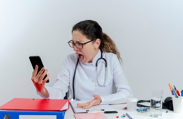 Jovem médica zangada usando túnica médica, estetoscópio e óculos, sentada na mesa com ferramentas médicas, segurando olhando para o telefone celular isolado