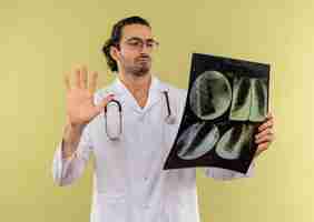 Foto grátis jovem médico a pensar com óculos ópticos, vestindo túnica branca com estetoscópio segurando e olhando para o raio-x mostrando gesto de parada no verde