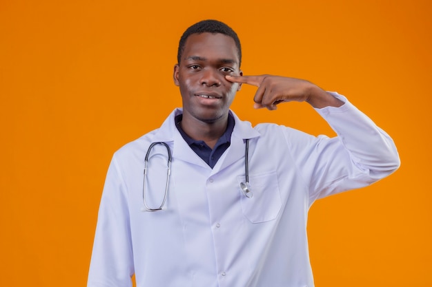 Jovem médico afro-americano vestindo jaleco branco com estetoscópio apontando o dedo para o olho, sorrindo e esperando