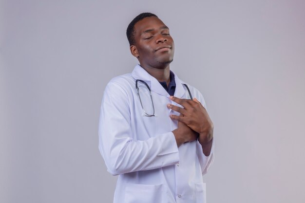 Jovem médico afro-americano vestindo jaleco branco com estetoscópio com os olhos fechados com as mãos cruzadas no peito, gesto de agradecimento sentimento positivo