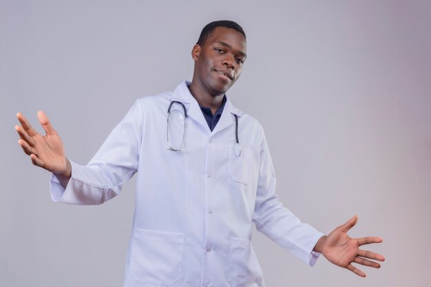 Jovem médico afro-americano vestindo jaleco branco com estetoscópio espalhando as palmas das mãos e abrindo as mãos fazendo um gesto de boas-vindas sorrindo