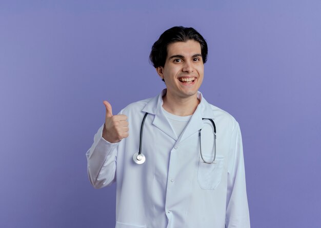 Jovem médico alegre vestindo túnica médica e estetoscópio mostrando o polegar isolado na parede roxa com espaço de cópia