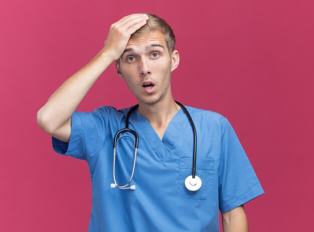 Jovem médico assustado vestindo uniforme de médico com estetoscópio colocando a mão na cabeça isolada na parede rosa