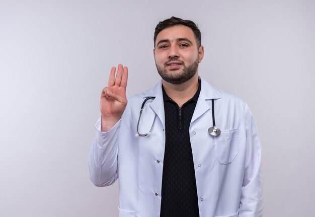Jovem médico barbudo vestindo jaleco branco com estetoscópio aparecendo e apontando para cima com os dedos número três sorrindo