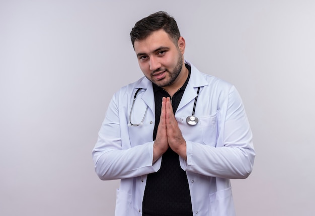 Jovem médico barbudo vestindo jaleco branco com estetoscópio de mãos dadas em gesto de oração, sentindo-se grato e feliz