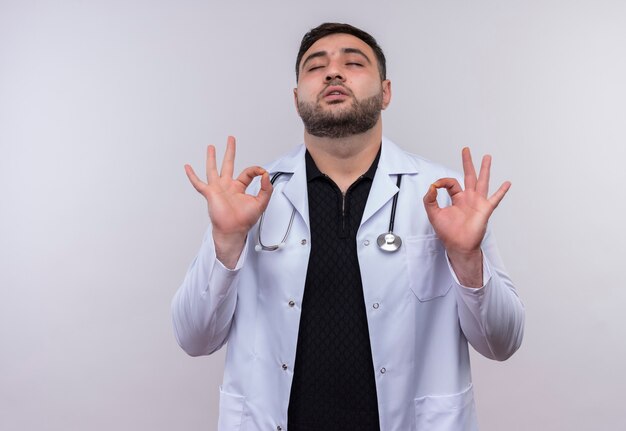 Jovem médico barbudo vestindo jaleco branco com estetoscópio e os olhos fechados relaxando fazendo sinais de meditação com os dedos