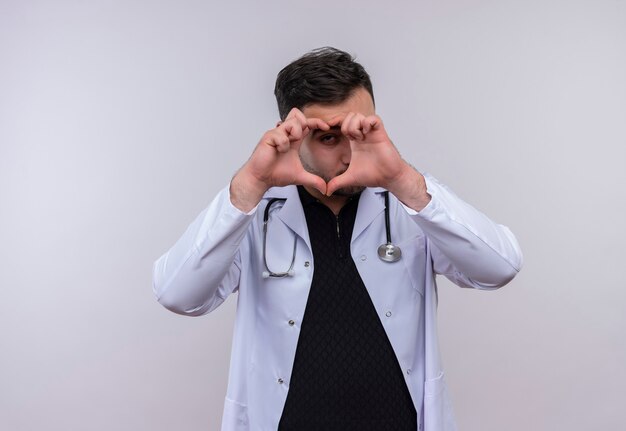Jovem médico barbudo vestindo jaleco branco com estetoscópio fazendo gestos de coração com os dedos olhando por entre os dedos