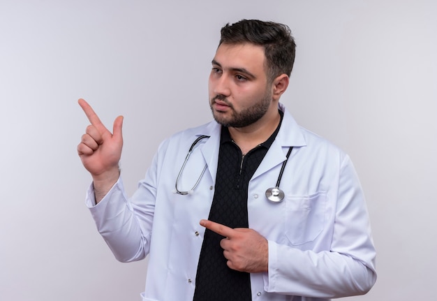Jovem médico barbudo vestindo jaleco branco com estetoscópio olhando para o lado com expressão séria e confiante apontando para o lado