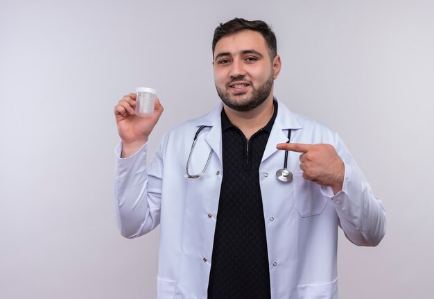Jovem médico barbudo vestindo jaleco branco com estetoscópio segurando o frasco de teste apontando com o dedo para ele sorrindo confiante